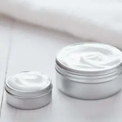 Dwa słoiczki kremu Nivea, jeden większy, drugi mniejszy, stoją na białym drewnianym blacie. Krem Nivea do czego? Do nawilżania i pielęgnacji skóry.