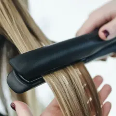 Dłoń trzyma prostownicę, wygładzając blond włosy. Nie wiadomo, ile stopni ma prostownica, ale efekt jest widoczny.