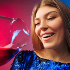 Uśmiechnięta kobieta w błyszczącej sukience, z kieliszkiem wina. Czy po botoksie można pić alkohol?