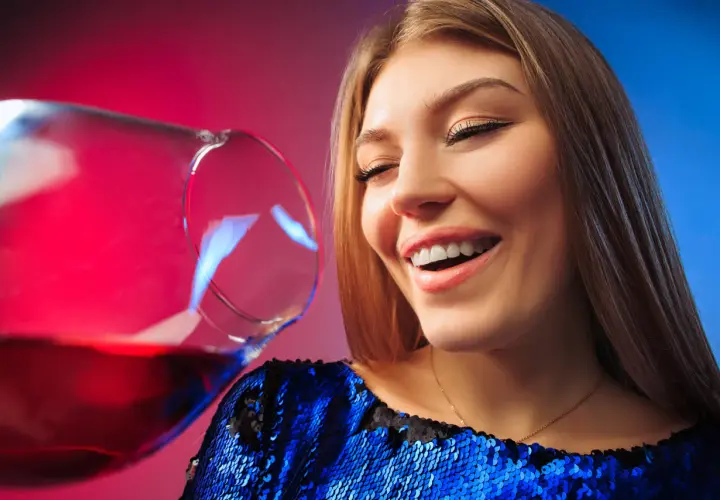 Uśmiechnięta kobieta w błyszczącej sukience, z kieliszkiem wina. Czy po botoksie można pić alkohol?