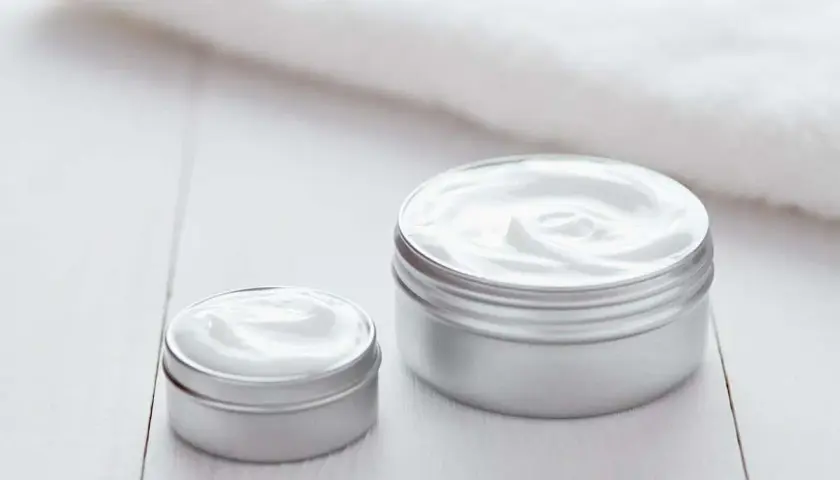 Dwa słoiczki kremu Nivea, jeden większy, drugi mniejszy, stoją na białym drewnianym blacie. Krem Nivea do czego? Do nawilżania i pielęgnacji skóry.