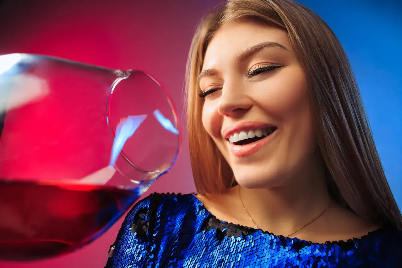 Uśmiechnięta kobieta w błyszczącej sukience, z kieliszkiem wina. Czy po botoksie można pić alkohol?
